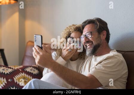 Happy couple in eyeglasses video chatting with close ones using mobile phone sitting on sofa at home. Joyful couple doing video call using mobile phon Stock Photo