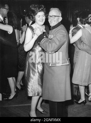 A young woman is dancing with an elderly ‘Chelsea Pensioner’ army veteran at the Zodiac Cocktail bar, Jermyn Street, London. Early 1960s. The gentleman is a resident at the Royal Hospital Chelsea, a retirement home and nursing home for former members of the British Army located in Chelsea, London. He is wearing his scarlet pensioner’s uniform coat and his military medals, which include First and Second World War campaign medals and a British Army Long Service and Good Conduct Medal. Stock Photo