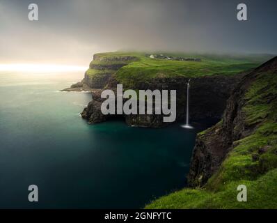 Gasadalur / Mulafossur Waterfall during a foggy/hazy sunset on the Faroe Islands on a lush green Summer day. Stock Photo