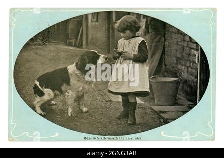 Original sentimental Edwardian greetings real photographic postcard, of a young Edwardian / Victorian girl in a farmyard wearing a pinafore feeding scraps to a waiting springer spaniel  - 'Hope deferred maketh the heart sick' is the inscription. posted April 1904, Published by Marcus Ward & Co. U.K. Stock Photo