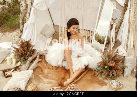 Young woman in white lace wedding dress in boho style sitting barefoot, on the beach. Boho style wedding decoration. Sea wedding concept. Stock Photo