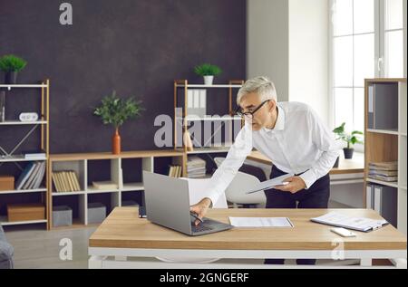 Senior busy businessman in the office checks statistics on a laptop and reads a paper report. Stock Photo