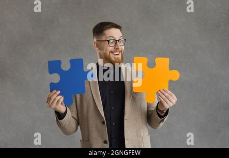 Funny smiling male office worker, makes up two colored puzzle pieces standing on a gray background. Stock Photo