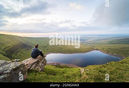 Man sits on a rock and looks at Llyn y Fan Fach lake. Brecon Beacons National Park. Black Mountain, Carmarthenshire, South Wales, the United Kingdom. Stock Photo