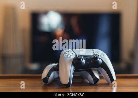 London, UK - May 25, 2021: Two Playstation 5 (PS5) controllers at home, tv on background, selective focus. Playstation 5 is the latest video game cons Stock Photo