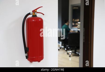 fire extinguisher without writing on office wall. Image of out of focus man working in office. Stock Photo