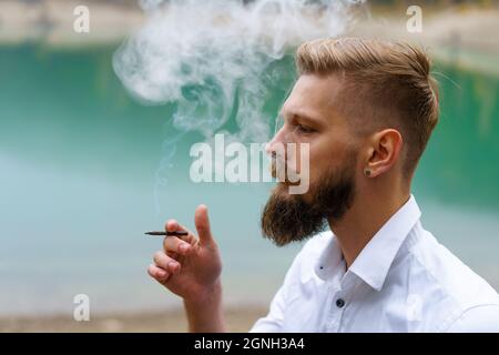 Young man beard smokes cigarette or tobacco. Guy is addicted to cigarettes and always smokes lot of tobacco every day. He smokes cigarette outdoors on background of lake in park. Cool guy blows smoke Stock Photo