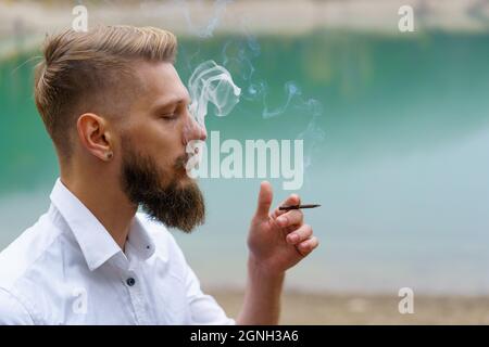 Business caucasian young bearded man in a white shirt smokes a cigarette and blows smoke against the backdrop of a turquoise lake in reflections. Faces solving problems and making choices Stock Photo