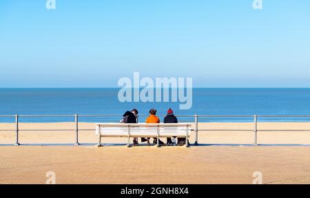 Waterfront promenade bench with three people sitting in front of Oostende (Ostend) North Sea beach, West Flanders, Belgium. Stock Photo
