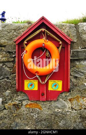 A bright orange lifesaving ring hangs on a red waterfront rescue station on the seawall surrounding the inner harbor at the village of Howth, Ireland. Stock Photo