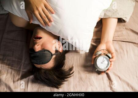 Hands of a young man from under the blankets hold an alarm clock in gray.  Stock Photo