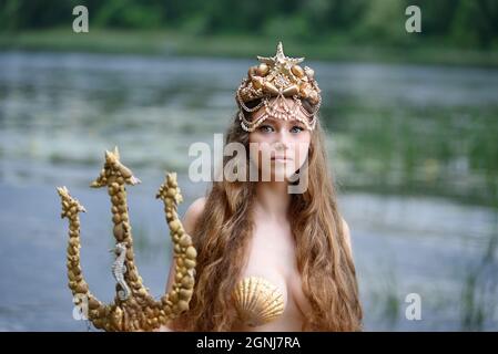 Fantasy woman real mermaid with trident myth goddess of sea with golden tail sitting in sunset on rocks.. Gold hair crown shells pearls jewelry. Merma Stock Photo