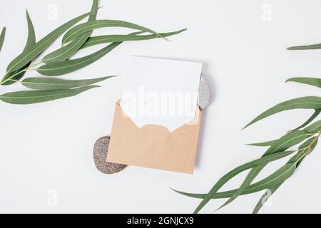 Mockup paper card in brown envelope on natural stones next to green eucalyptus branches on white background, top view Stock Photo