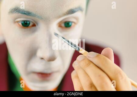 Mim is preparing makeup for performance. Close up of clown cosplayer. Guy with green hair in burgundy suit. Theatre atmosphere. Male actor is covering his face by white greasepaint using brush. Stock Photo
