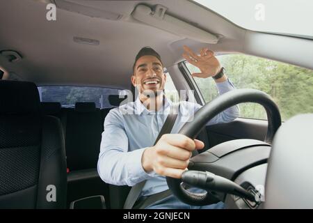 Cheerful middle-eastern guy driving auto, greeting someone