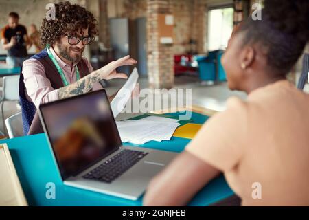young caucasian beardy guy with glasses and tattoo handing over a document to his afro-american female colleague Stock Photo