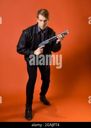 a fighter for justice, a guy in a casual shirt and a leather jacket with a shotgun, ready to attack, studio photo on an orange background Stock Photo