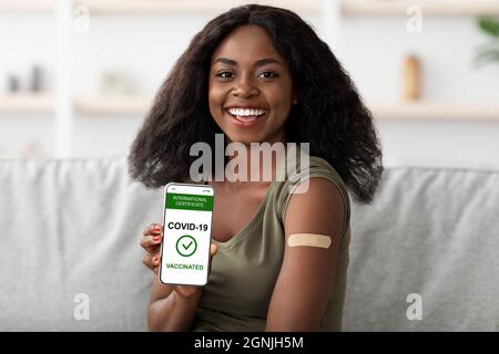 Vaccinated african-american woman showing cellphone with vaccination certificate, home inetrior Stock Photo