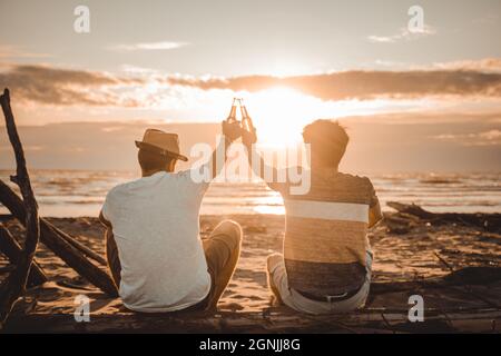Two young friends spending time together sitting on the beach, drinking beer and toasting on vacation in twilight summer sunset. Friendship concept Stock Photo
