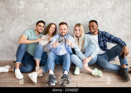 Group of young diverse friends using modern smartphone together, sitting near grey studio wall, full length Stock Photo