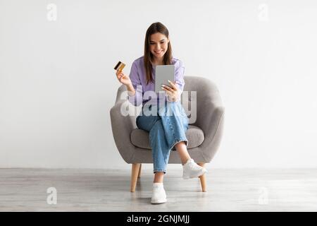 Cool young lady with digital tablet and credit card shopping online, sitting in armchair against white studio wall Stock Photo