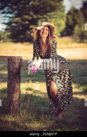 Young woman in summer dress and hat leaning on the wooden fence. Countryside Stock Photo