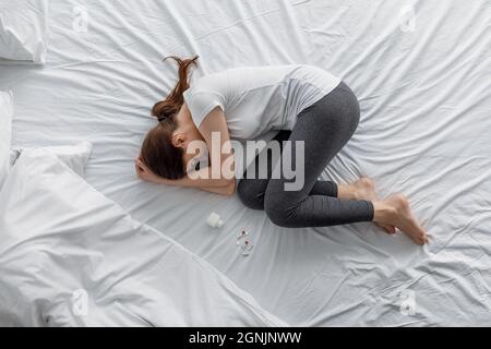 Sad despair young caucasian woman suffering from abdominal pain lying on bed with jar and scattered pills Stock Photo