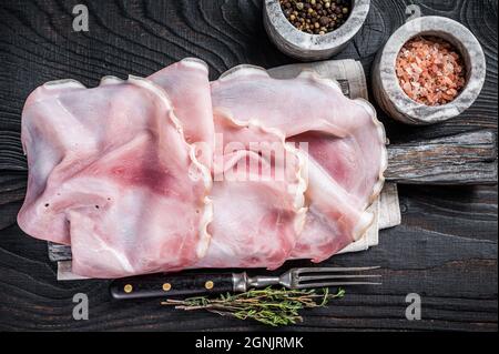 Sliced prosciutto ham on wooden board with herbs. Black wooden background. Top view Stock Photo