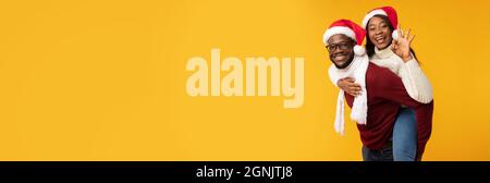 Black Man Carrying Woman Piggyback Wearing Santa Hats, Yellow Background Stock Photo