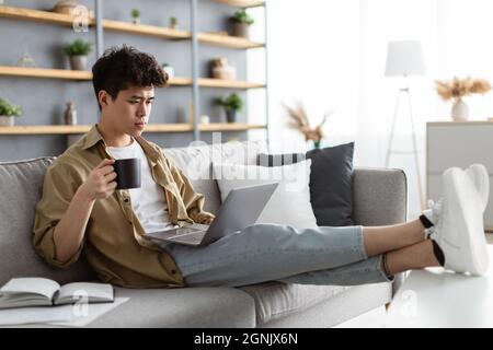 Serious asian man watching video on pc, drinking hot coffee Stock Photo