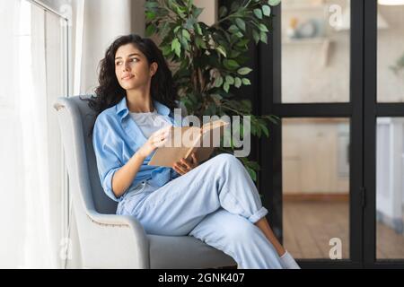Thoughtful young woman sitting in comfy armchair with open book, looking out window, daydreaming indoors, copy space Stock Photo