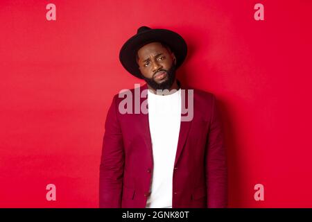 Sad and unamused Black man looking with pity at camera, frowning disappointed, standing against red background Stock Photo