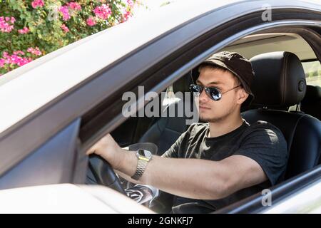 a teenager is driving. a modern young guy with sunglasses, fashionable with a beard, drives a white car. driving training or car travel. Stock Photo