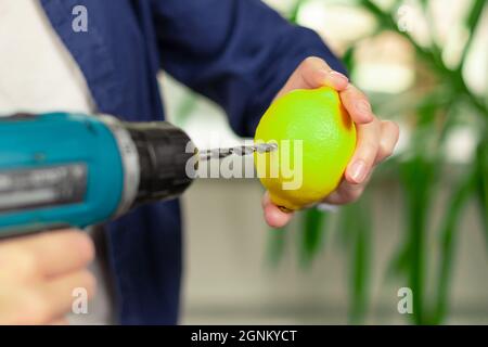 Female hands in a blue shirt hold a screwdriver with a drill and a bright lemon against the background of a light window and green leaves of a housepl Stock Photo