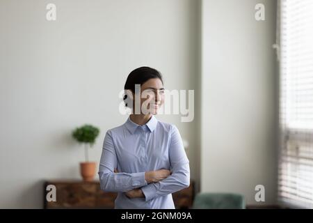 Happy millennial Indian businesswoman looking at window with folded arms Stock Photo