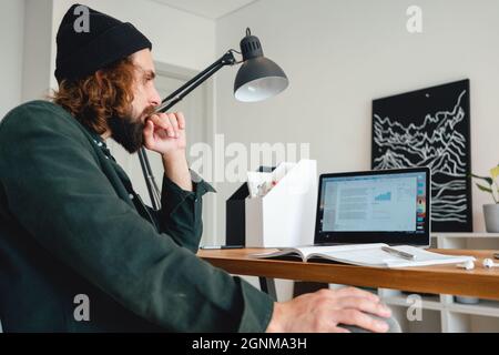 Bearded college student preparing for pass exam at home using laptop. Young man making notes while online learning Stock Photo