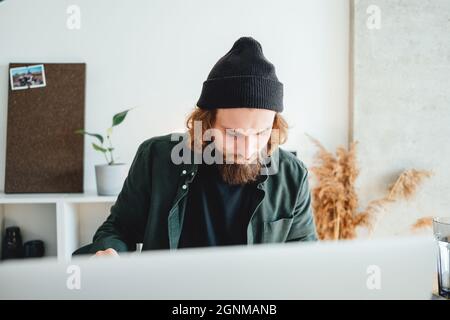 Bearded college student preparing for pass exam at home using laptop. Young man making notes while online learning Stock Photo