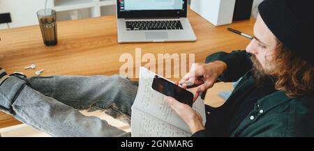Bearded college student preparing for pass exam at home using smartphone. Young man making notes while online learning. Wide image Stock Photo