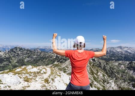 Woman celebrating mountaineering achievement with arms raised in the air, while looking at beautiful mountaintops landscape Stock Photo
