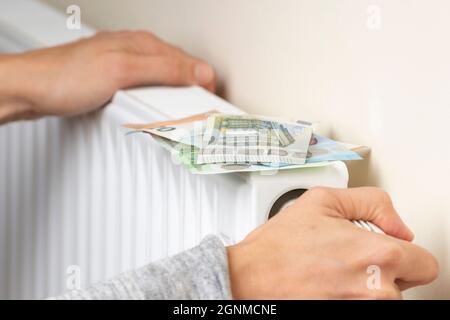 Female hand checking and adjusting the temperature of heating radiator at home. Euro money banknotes on heating battery. Expensive heating costs Stock Photo