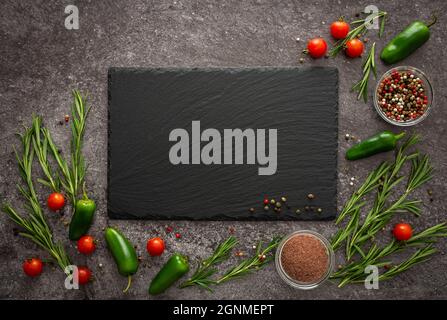 Black empty rectangular slate board on gray background surrounded by fresh cherry tomatoes, jalapeno peppers, spices and rosemary Stock Photo
