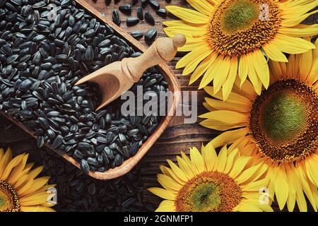 Wooden bowl of black sunflower seeds, beautiful yellow sunflowers on wooden board, top view, flat lay. Stock Photo