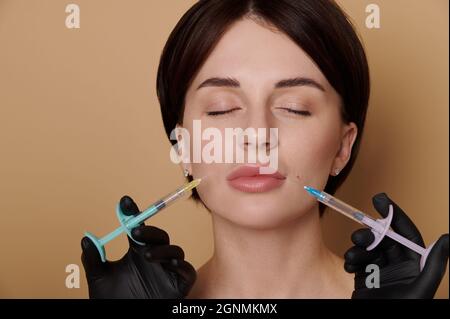 Headshot of beautiful young woman receiving beauty injections on her face. Hands in black medical gloves holding syringes with hyaluronic acid product Stock Photo