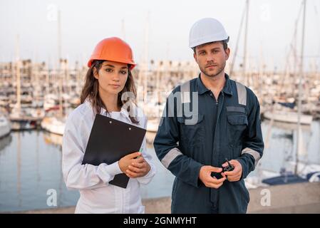 Professional young male and female marine colleagues in hardhats and workwear with clipboard and VHF radio looking at camera while having meeting on pier and inspecting sea port Stock Photo