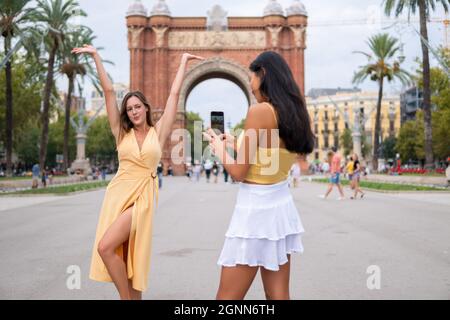 Young woman in stylish yellow dress posing with arms raised against historical triumphal arch while Asian girlfriend photographing on smartphone during sightseeing in Barcelona city in summer day Stock Photo
