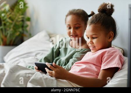 Portrait of two cute girls using smartphone together while sitting lying on bed in cozy room and watching videos Stock Photo