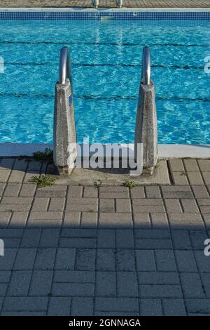 A view from behind a ladder at the edge of an outdoor public pool shows its railings supported by concrete. Stock Photo