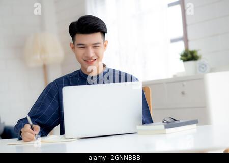 Young asian business man writing on notebook for planning working and using laptop computer on desk at home, notes about finance, male study and learn Stock Photo