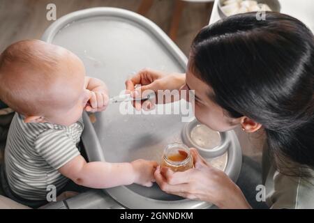 First baby food pure. Mother giving spoon of vegetables or fruits in feeding chair at home. Cute child trying to eat by himself. healthy support Stock Photo