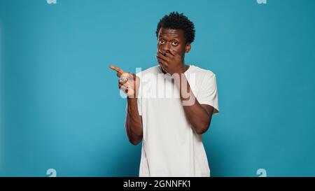 Portrait of person covering mouth with hands after telling secret by mistake. Young man talking about rumour and gossip by accident. Adult ruining surprise and pointing at left side Stock Photo